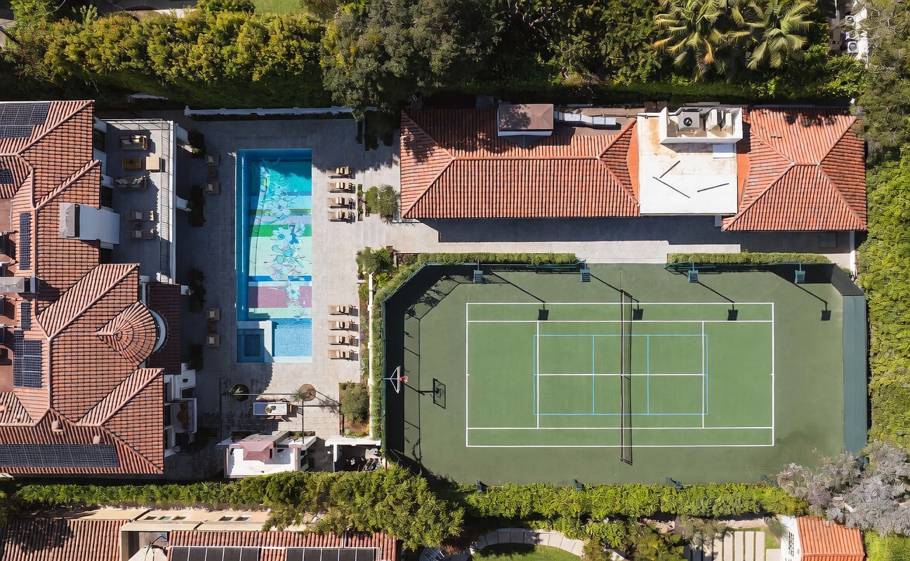 This high-angle aerial view captures a luxurious residential property featuring a large, rectangular swimming pool with a decorative mosaic floor and an adjacent full-sized tennis court. The grounds include a spacious paved patio area, a long single-story structure with a terracotta-tiled roof, and lush perimeter landscaping that provides significant privacy. The perspective emphasizes the expansive outdoor recreational amenities and the well-organized layout of the estate.