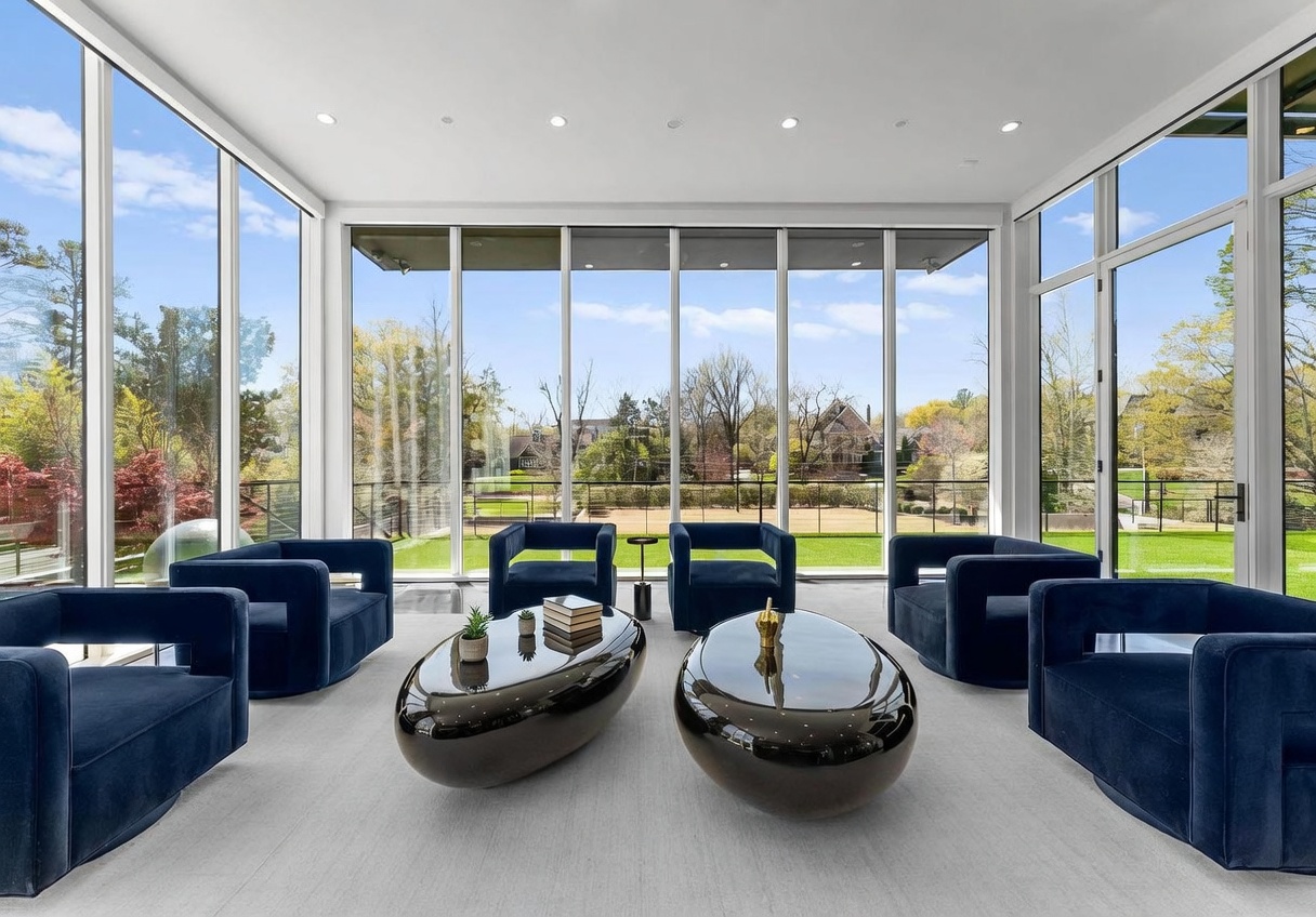 This bright and airy sunroom features expansive floor-to-ceiling windows that provide an unobstructed view of the lush outdoor landscape. The space is furnished with four plush, deep-blue velvet armchairs arranged around two sleek, dark, oval-shaped coffee tables. The minimalist design and neutral carpeting create a sophisticated, tranquil atmosphere perfect for relaxation or conversation.