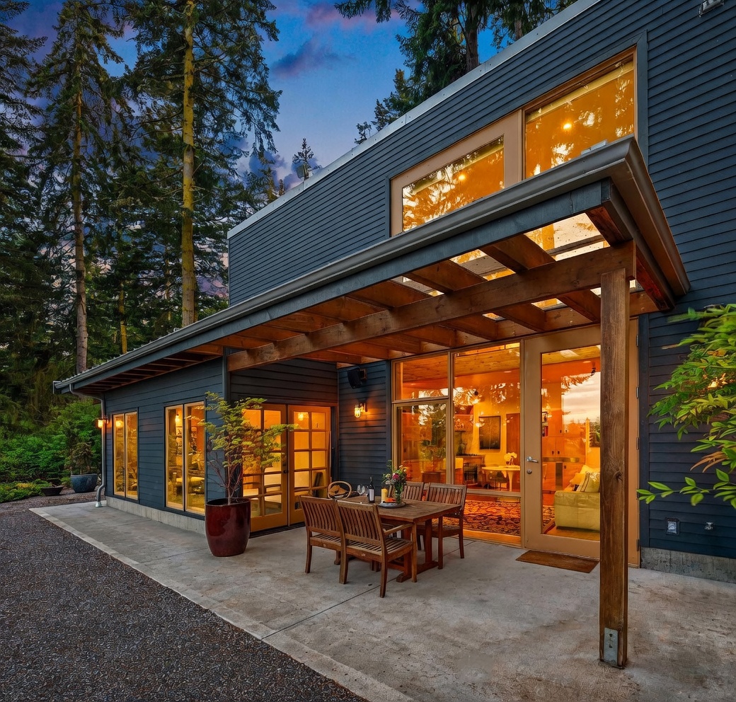 This image captures a modern, dark-sided home featuring a spacious concrete patio area perfect for outdoor dining. A wooden pergola extends from the house, providing partial shade over a dining set, while large glass doors and windows offer a seamless transition between the indoor living space and the exterior. The setting is framed by tall, lush trees under a twilight sky, creating a serene and inviting atmosphere.