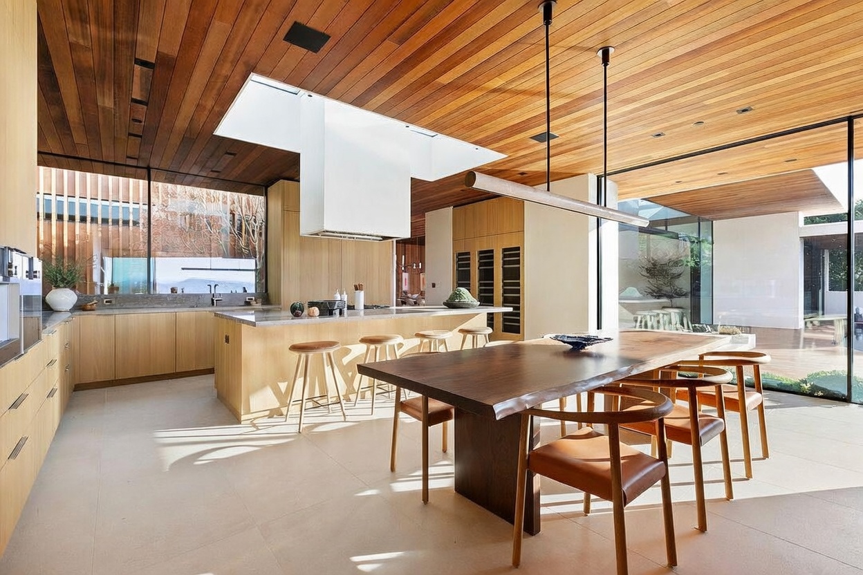 This stunning, modern kitchen features a seamless blend of natural wood ceilings and light-toned cabinetry, creating a warm and sophisticated atmosphere. A large, live-edge wooden dining table sits in the foreground, complemented by a spacious kitchen island with bar seating and a prominent white range hood. The open-concept design is enhanced by floor-to-ceiling glass walls that invite abundant natural light and provide a clear view of the outdoor landscape.