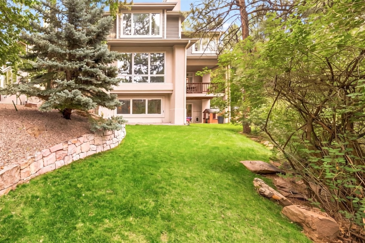 This rear view of a multi-story home showcases a well-maintained lawn bordered by a stone retaining wall and mature trees. The house features a neutral-toned exterior with large windows across multiple levels and a covered wooden deck on the middle floor. The setting is lush and wooded, providing a private and serene atmosphere.