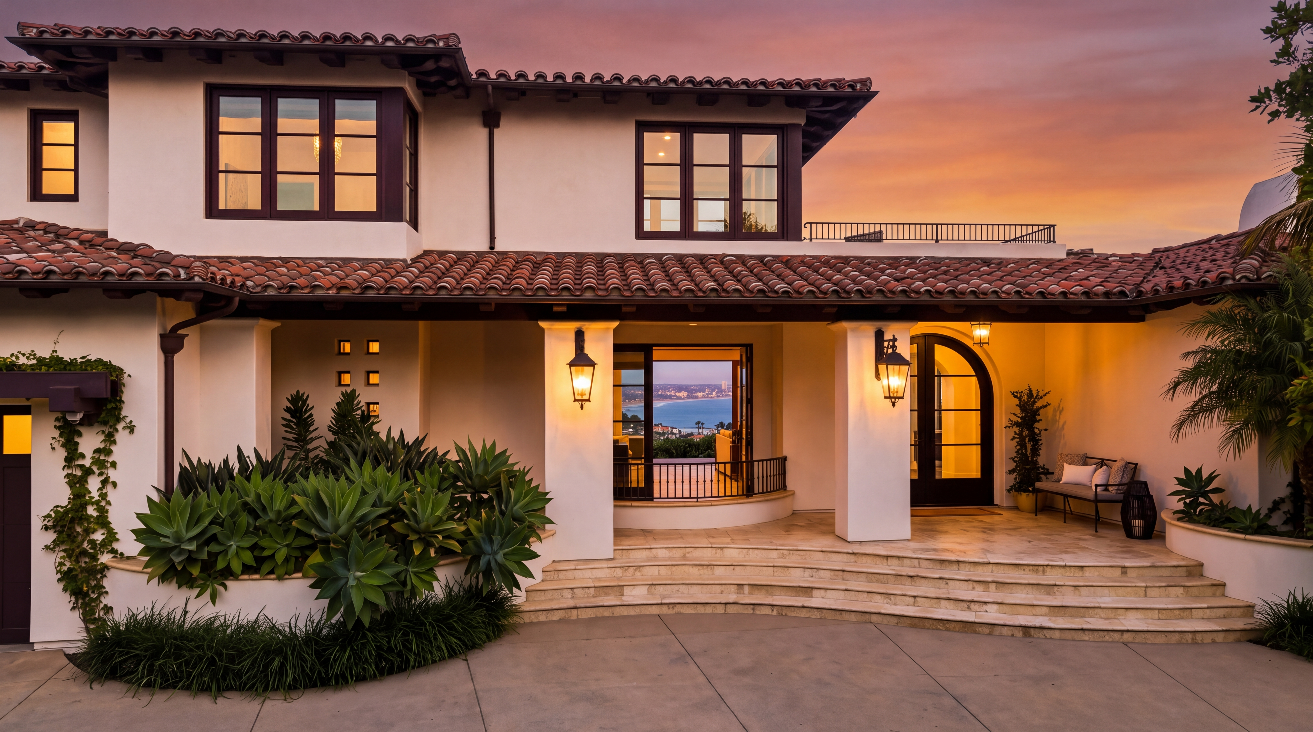This elegant Mediterranean-style home features a grand front entryway with a covered porch, stone steps, and warm ambient lighting. The exterior is characterized by white stucco, a tiled roof, and lush landscaping, while the open door frames a picturesque sunset view of the coastline. The architectural design creates a sophisticated, welcoming impression perfect for luxury living.