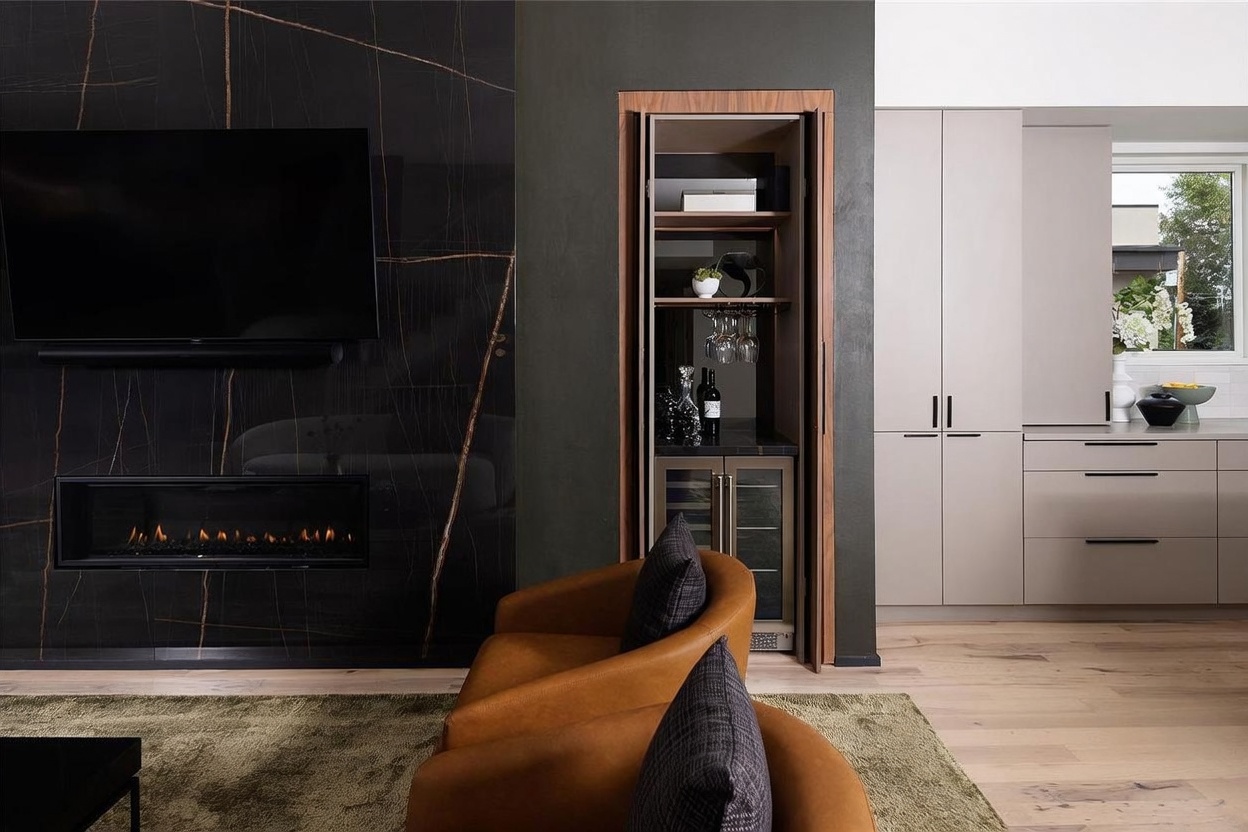 This contemporary living space features a striking black stone-clad wall with integrated fireplace and television. To the right, a built-in bar cabinet with glass shelving and a wine cooler is recessed into the wall, adjacent to a sleek kitchen area with light-toned cabinetry. Two warm-toned leather armchairs sit on a textured rug, creating a sophisticated and inviting atmosphere.