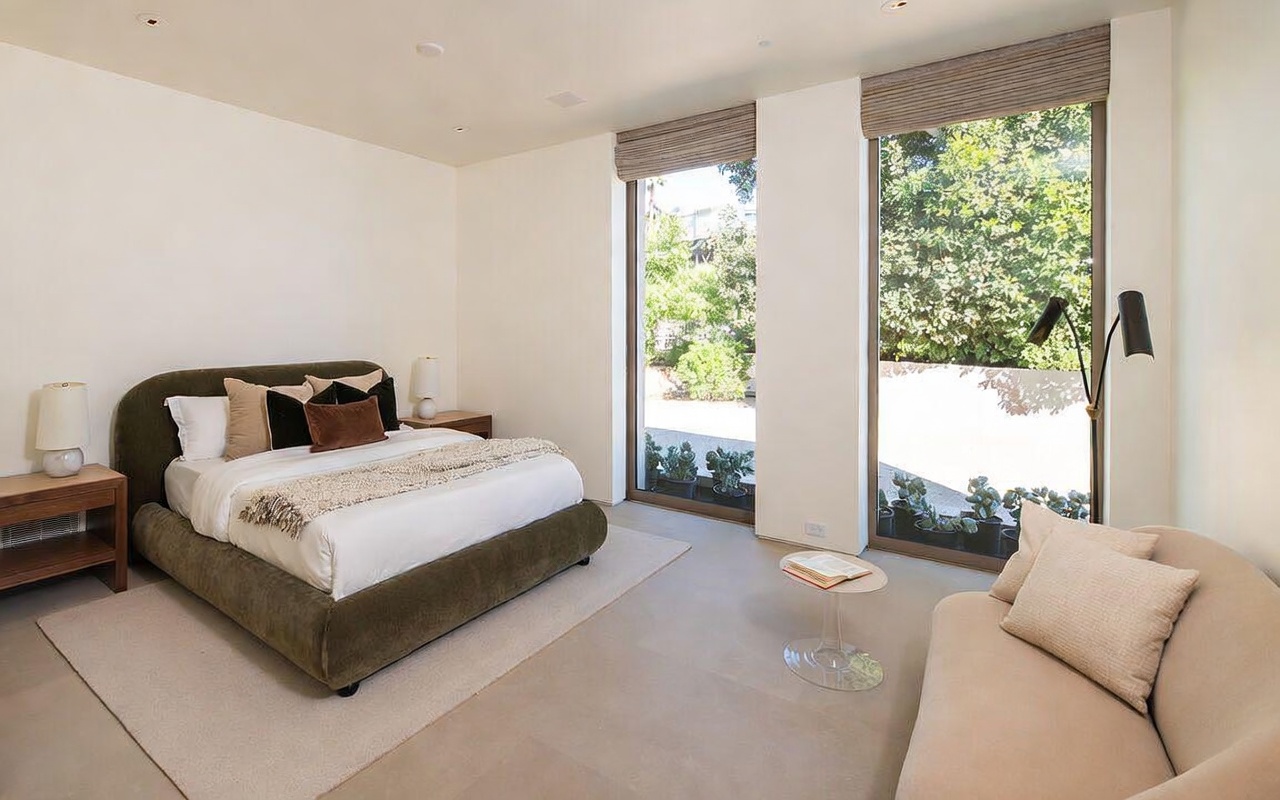 This serene primary bedroom features a plush, olive-green upholstered bed as the focal point, complemented by minimalist bedside tables and lamps. Large floor-to-ceiling windows flood the room with natural light, offering a view of the lush greenery outside, while a sleek beige sofa provides a cozy seating area. The neutral color palette and clean lines create a sophisticated, tranquil atmosphere perfect for relaxation.