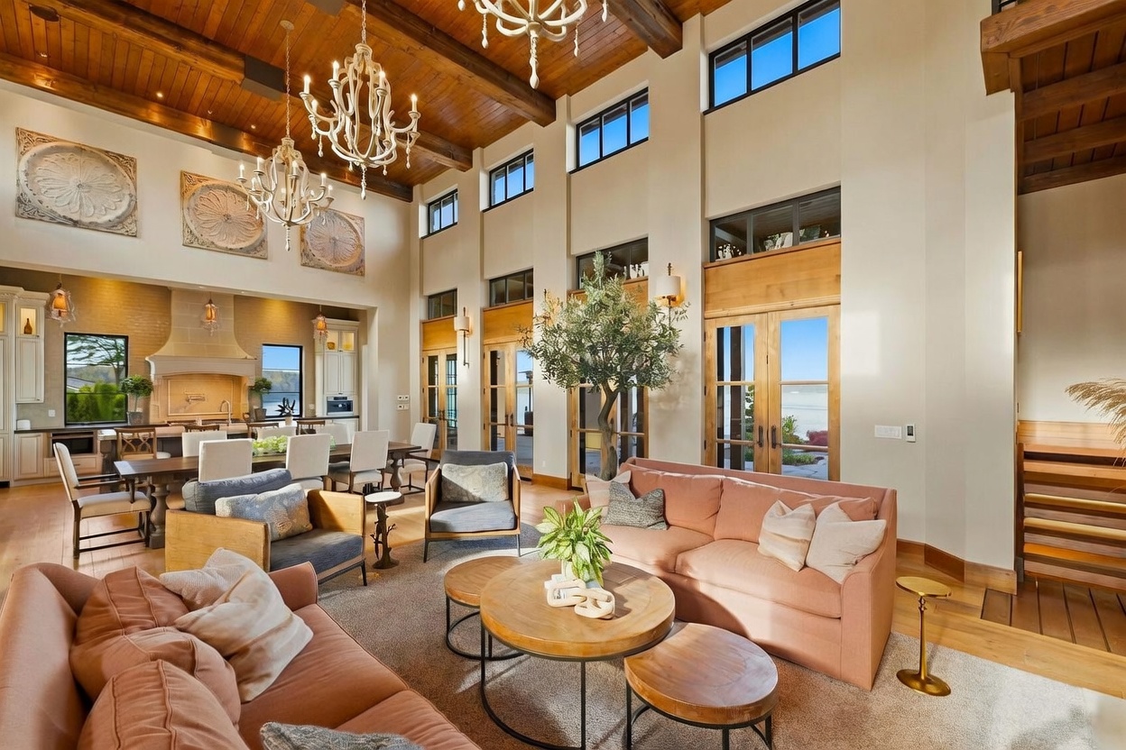 This expansive, open-concept living area features a vaulted wood-paneled ceiling with exposed beams and elegant chandeliers. The space is anchored by a comfortable seating arrangement with plush sofas and armchairs centered around a tiered wooden coffee table, while a large indoor tree adds a natural touch. Large windows and glass doors provide abundant natural light and views of the outdoors, seamlessly connecting the living room to the adjacent dining and kitchen area.