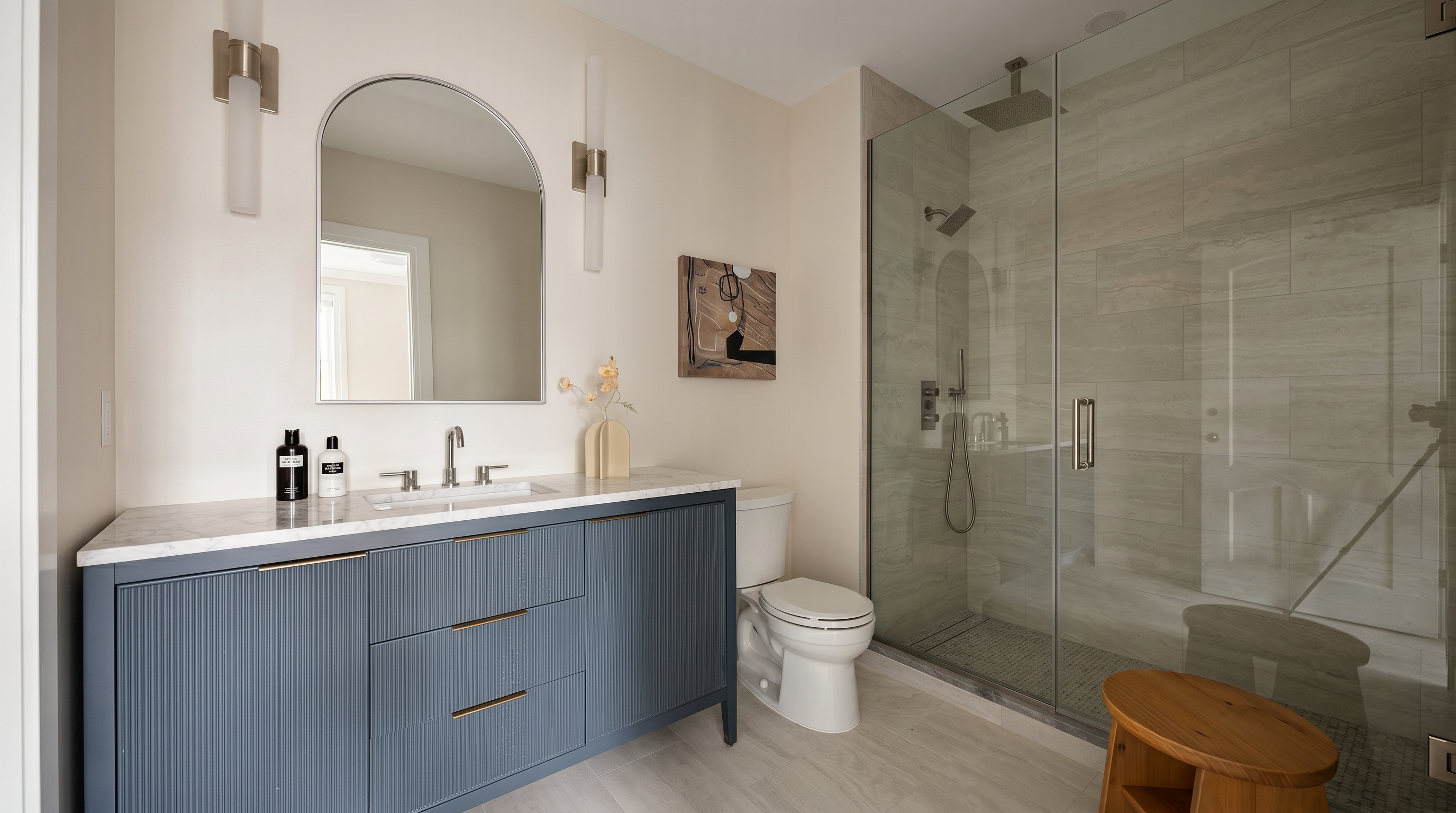 This modern primary bathroom features a striking blue fluted vanity with gold hardware, topped with a marble countertop and an arched mirror. To the right, a spacious walk-in shower with glass doors and neutral stone-look tile creates a clean, spa-like aesthetic. The room is bright and airy, accented by contemporary wall sconces and a small wooden stool, offering a sophisticated and tranquil atmosphere.