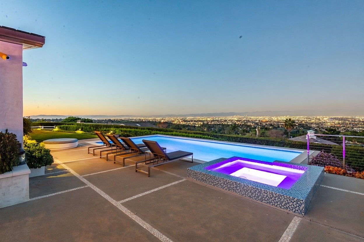 This stunning outdoor space features a rectangular infinity-edge pool and a raised, tiled spa with vibrant purple underwater lighting. A row of lounge chairs is arranged on the concrete patio, which overlooks a sprawling panoramic view of the city lights below at dusk. The scene is framed by manicured hedges and a clear, expansive sky, creating a luxurious and serene atmosphere.