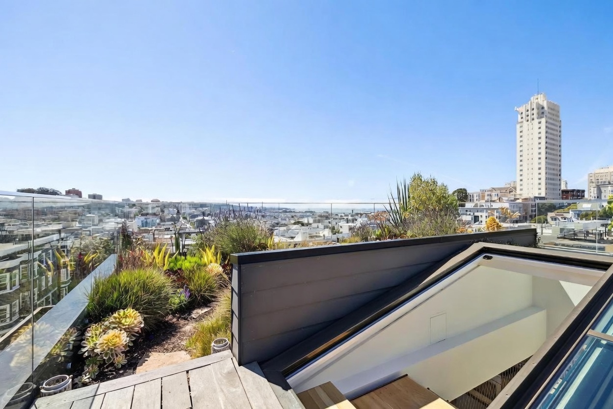 This rooftop deck offers a stunning panoramic view of the city skyline and the bay under a clear blue sky. The space features a modern glass railing, a landscaped garden bed with various succulents and grasses, and a wooden deck surface. A skylight structure provides a glimpse into the interior, highlighting the seamless transition between indoor and outdoor living spaces.