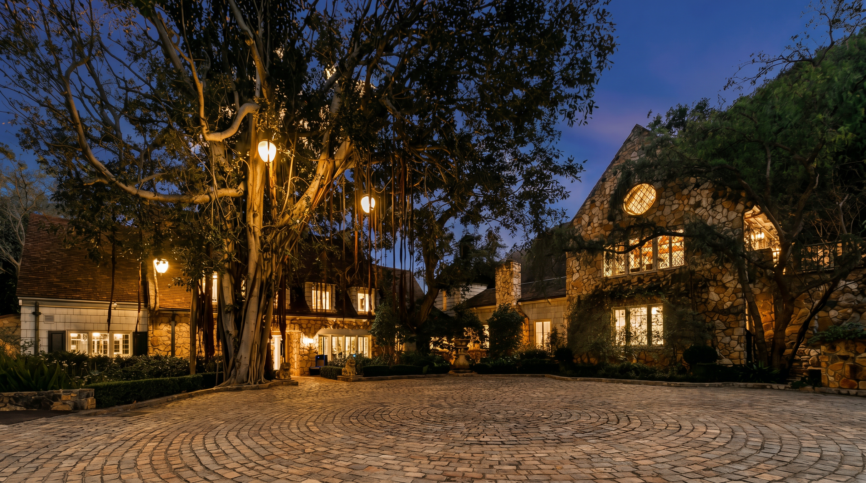 This stunning estate features a grand, stone-clad exterior with a classic European architectural style, illuminated beautifully against a twilight sky. A large, circular cobblestone driveway leads up to the main entrance, framed by mature trees and lush landscaping. The scene captures a sense of timeless elegance and luxury, perfect for a high-end property listing.