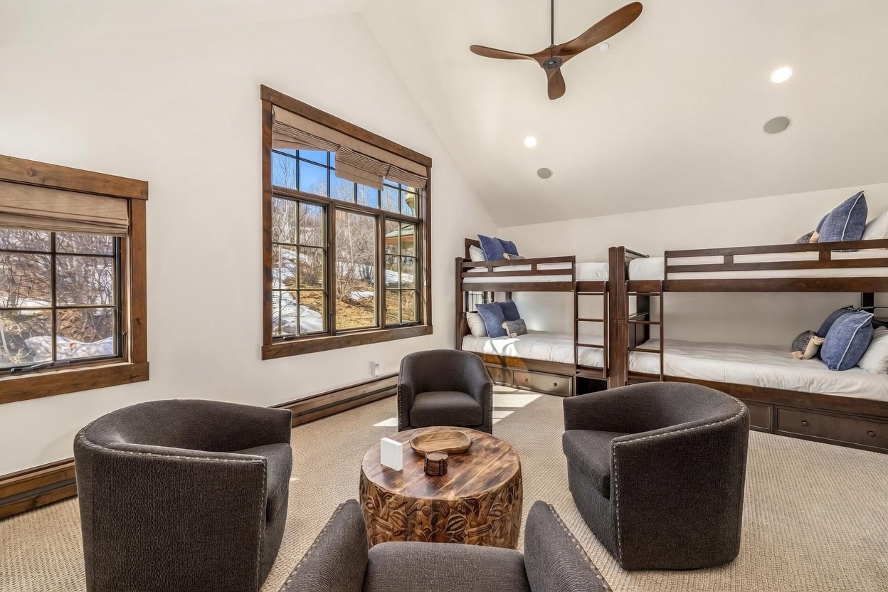 This spacious bedroom features a vaulted ceiling with a wooden fan and two sets of dark wood bunk beds, providing ample sleeping space. A cozy seating area is arranged in the center of the room with four dark upholstered armchairs surrounding a rustic, carved wooden coffee table. Large windows with wooden trim offer views of the snowy landscape outside, creating a warm and inviting mountain-style retreat.