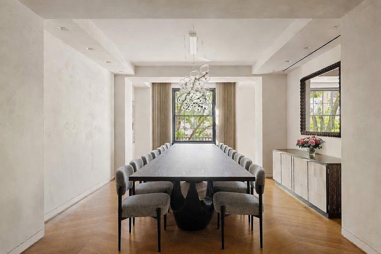 This elegant dining room features a long, dark-toned table surrounded by twelve upholstered chairs, centered beneath a striking, multi-globe glass chandelier. The space is anchored by light wood herringbone flooring and a long sideboard topped with a floral arrangement and a large, framed mirror. The room is framed by a window looking out onto greenery, creating a sophisticated and symmetrical atmosphere perfect for formal entertaining.