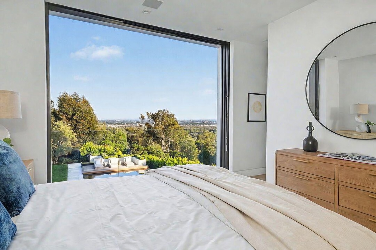 This bright and airy primary bedroom features a large, floor-to-ceiling window that frames a panoramic view of the surrounding landscape and a private outdoor deck. The room is furnished with a plush bed covered in white linens and a light wood dresser topped with a large circular mirror. The minimalist design emphasizes the connection between the indoor living space and the natural scenery outside.