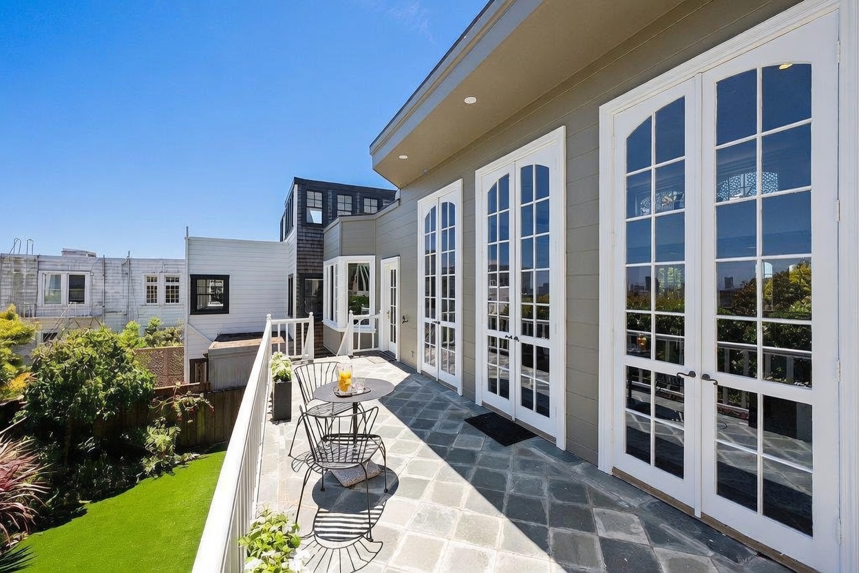 This elevated outdoor terrace features a stone-tiled floor and a white railing overlooking a lush green backyard. Multiple sets of tall glass-paned doors provide seamless indoor-outdoor access, while a small bistro set offers a cozy seating area. The perspective captures the bright, sunny atmosphere of the deck against a clear blue sky, highlighting the home's connection to the exterior space.