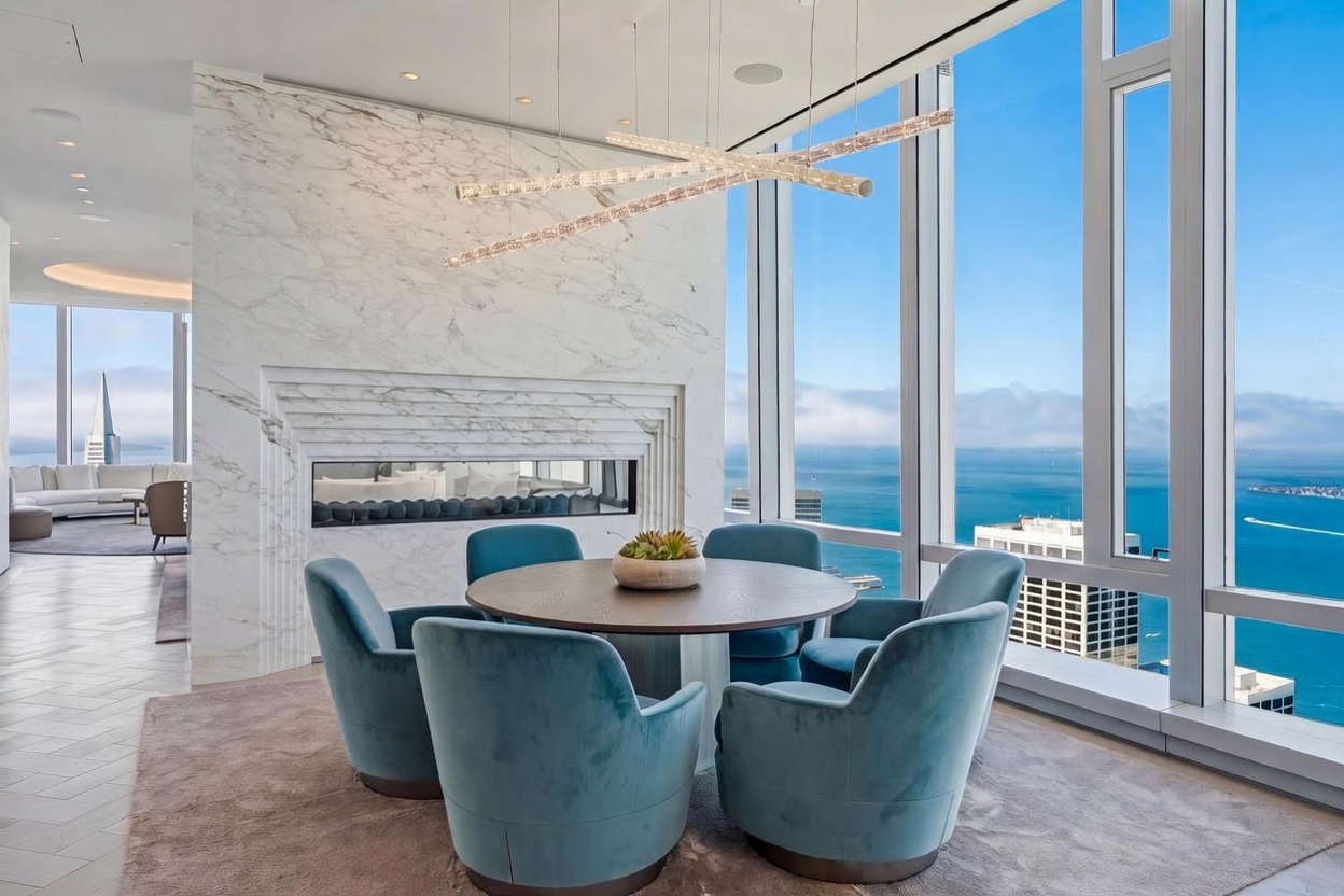 This sophisticated dining area features a round table surrounded by six plush, blue velvet chairs, set against a backdrop of expansive windows that offer a panoramic view of the bay and city skyline. A striking, multi-tiered linear light fixture hangs above the table, while a prominent stone-clad chimney breast with a built-in horizontal fireplace serves as a dramatic focal point. The space is defined by a neutral-toned area rug and a seamless transition into an adjacent living area, creating an open and airy atmosphere.