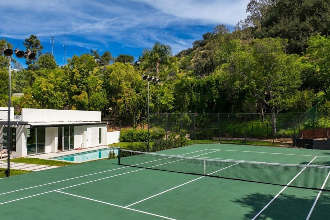 This outdoor space features a full-sized tennis court with a green surface, enclosed by high chain-link fencing. Adjacent to the court is a modern, white-walled structure with large glass doors and a small swimming pool, set against a backdrop of lush green hills and trees. The perspective is from the edge of the tennis court, capturing a bright, sunny day that highlights the recreational amenities of the property.