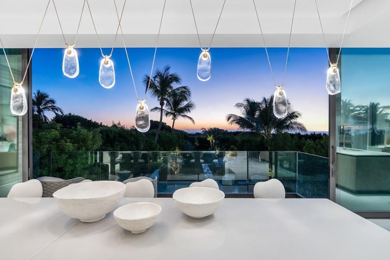 This sophisticated dining area features a sleek, minimalist table adorned with textured, organic-shaped bowls. Above, a series of unique, teardrop-shaped glass pendant lights hang at varying heights, creating a dramatic focal point. The room opens up through expansive glass walls to a stunning sunset view over a lush landscape and the distant ocean, blending indoor luxury with the natural environment.
