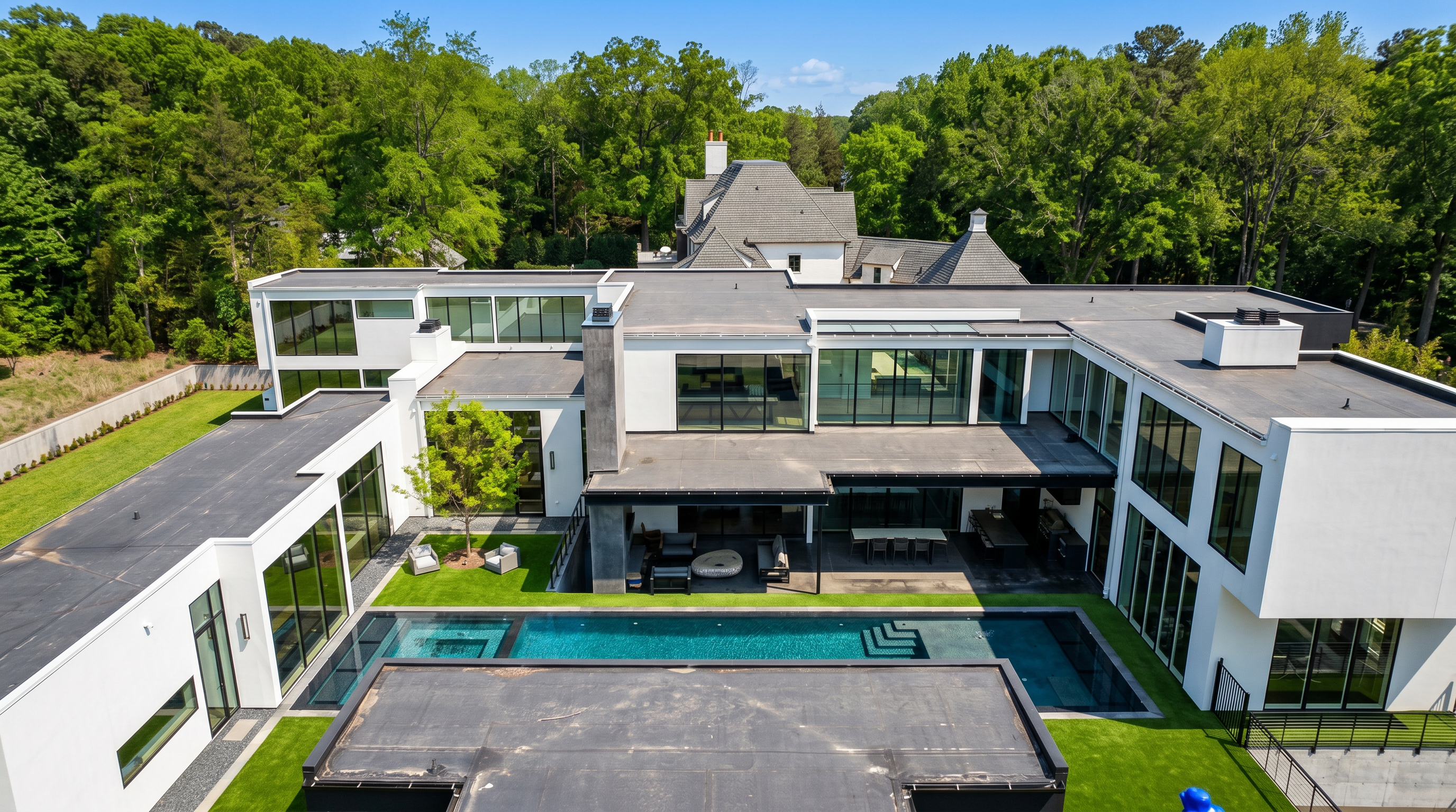 This high-angle aerial perspective captures a sprawling, modern luxury estate characterized by its clean lines, white exterior walls, and extensive use of glass. The property features a central courtyard with a manicured lawn and a tree, flanked by a large rectangular swimming pool and a covered outdoor living area. The architectural design emphasizes indoor-outdoor flow, with multiple wings of the house surrounding the private pool deck.