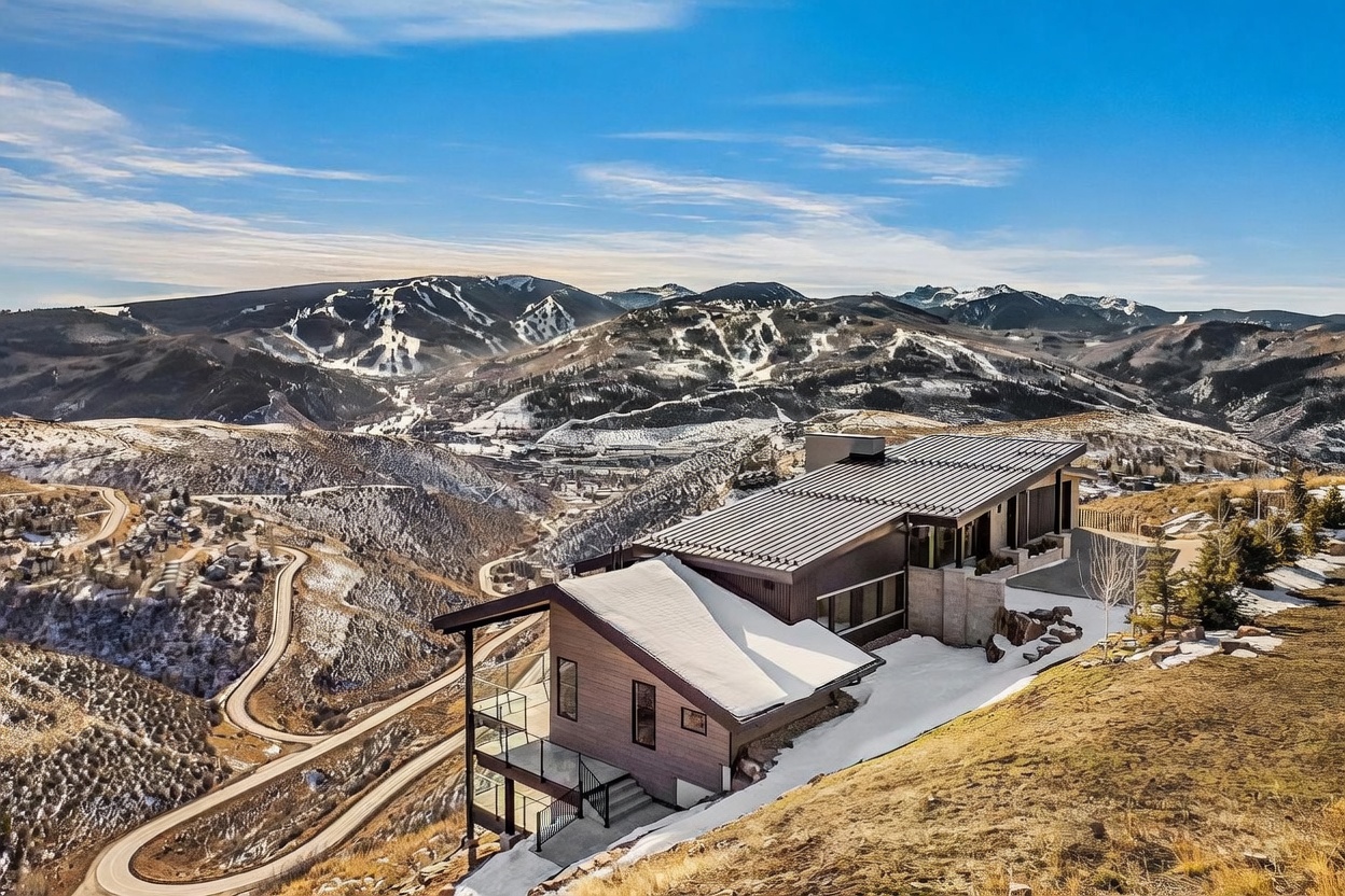 This aerial view showcases a modern mountain home nestled on a hillside, with stunning panoramic views of snow-capped mountains and winding roads. The house features a contemporary design with a metal roof, clean lines, and multiple levels, blending seamlessly with the natural landscape. The surrounding terrain is a mix of grassy slopes and patches of snow, emphasizing the property's unique location and access to outdoor recreation.