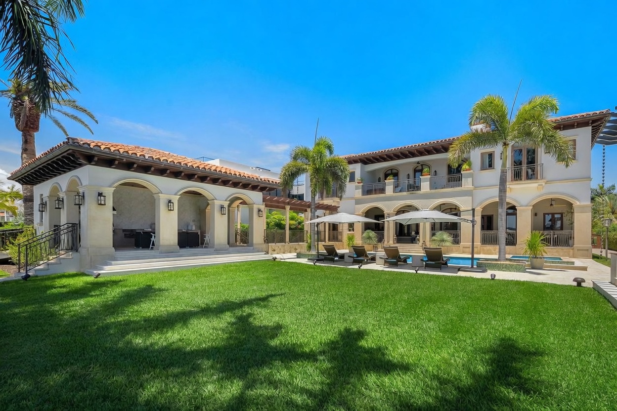 This expansive rear view of a luxury Mediterranean-style estate showcases a sprawling, manicured lawn leading to a multi-level residence with a private swimming pool. The property features a covered outdoor kitchen and lounge area, elegant arched architectural details, and multiple balconies overlooking the lush backyard. The bright, sunny atmosphere and clear blue sky emphasize the high-end, resort-like appeal of this outdoor living space.