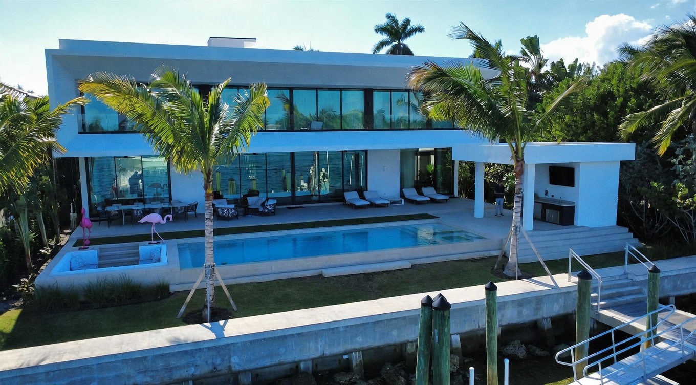 This modern waterfront estate features a sleek, two-story architectural design with extensive glass walls that offer seamless indoor-outdoor living. The rear exterior showcases a rectangular swimming pool, a spacious paved patio area with lounge seating, and a covered outdoor kitchen space. Lush tropical landscaping, including tall palm trees, frames the property, which overlooks a private dock and waterway.