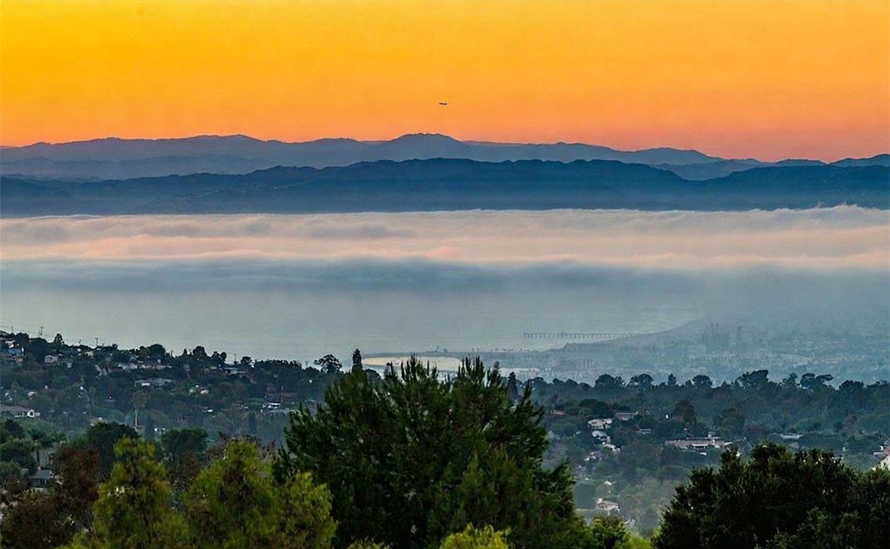 This scenic landscape view captures a breathtaking sunset over a coastal region, with a thick layer of low-hanging marine layer clouds obscuring the lower elevations. In the foreground, lush green trees frame the vista, while the distant mountains are silhouetted against a vibrant, warm-toned sky. The perspective is elevated, providing a panoramic outlook that emphasizes the property's prime location and expansive natural surroundings.