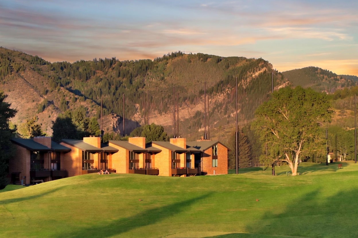This image features a multi-unit brick residential building set directly against a sprawling, well-maintained golf course fairway. The architecture is characterized by a series of repeating gabled rooflines, brick facades, and private balconies that overlook the lush green grass and the majestic mountain range in the background. The serene atmosphere and picturesque natural setting make this property an ideal location for outdoor enthusiasts and golf lovers.
