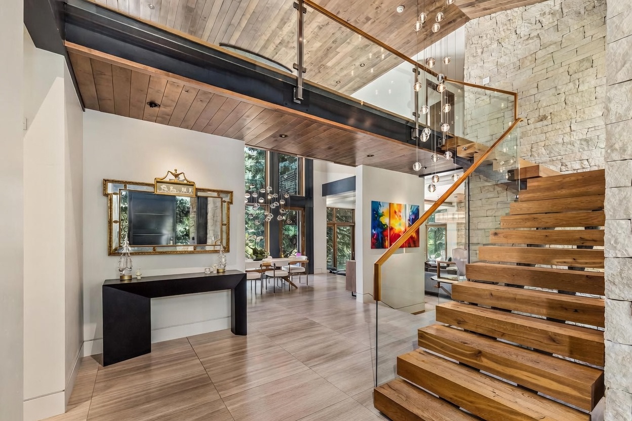 This grand entryway features a striking floating staircase with thick wood treads and a glass railing, set against a textured stone wall. A sleek black console table sits beneath an ornate gold-framed mirror, while a dramatic multi-pendant light fixture hangs from the wood-paneled ceiling. The open-concept design provides a clear line of sight through to the dining area, emphasizing the home's spacious and modern architectural aesthetic.
