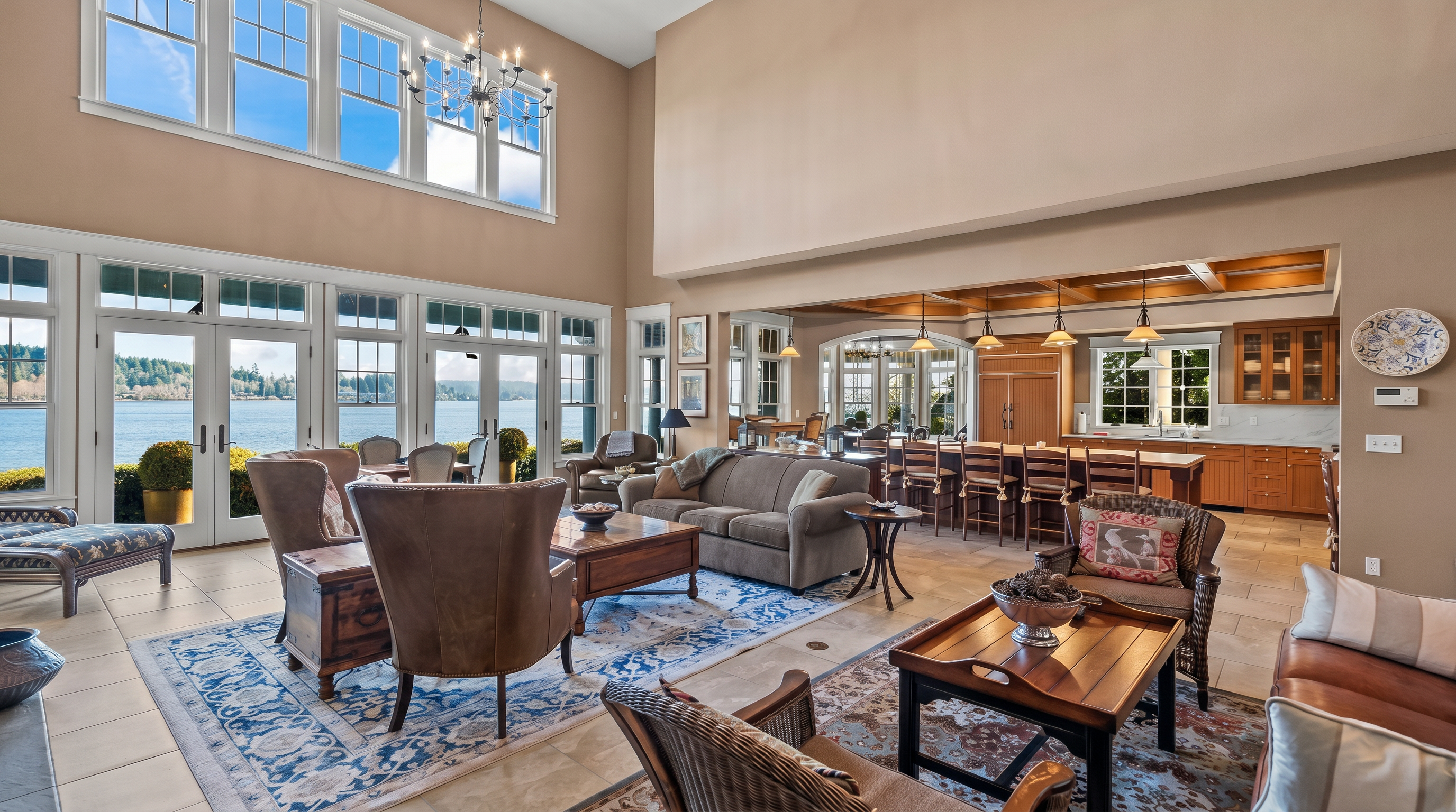 This expansive, open-concept living area features a soaring vaulted ceiling and a wall of large windows that provide panoramic water views. The space is furnished with comfortable seating arrangements, including plush sofas and armchairs set atop patterned area rugs, creating a warm and inviting atmosphere. Beyond the living area, a spacious kitchen with a long island and bar seating is visible, highlighting the home's seamless flow and suitability for entertaining.