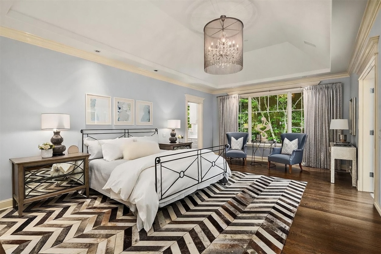 This spacious primary bedroom features a light blue color palette, a metal-framed bed, and a striking chevron-patterned area rug that anchors the room. A large drum-style chandelier hangs from the tray ceiling, while two blue armchairs are positioned near the large windows that overlook lush greenery. The room is completed by mirrored nightstands and a decorative console table, creating an elegant and serene atmosphere.