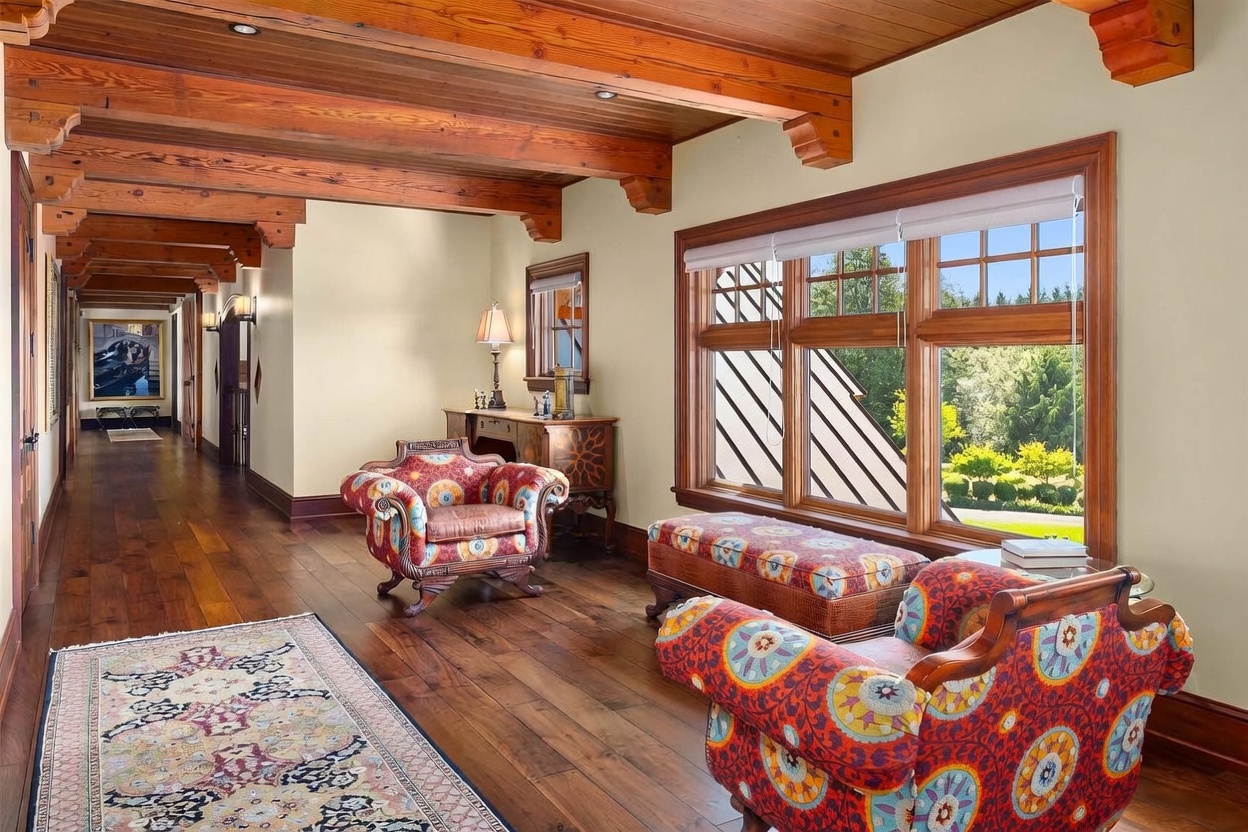 This elegant hallway features rich, dark wood flooring and a striking exposed beam ceiling that adds architectural character. The space is furnished with two patterned armchairs and a matching ottoman, creating a cozy seating area bathed in natural light from a large window. A decorative wooden console table sits against the wall, complemented by a traditional area rug that anchors the space.