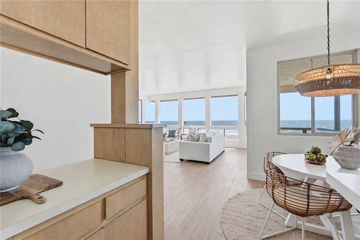 This bright, open-concept space captures a view from the dining area toward a living room that overlooks the ocean. The dining area features a round white table with woven chairs under a textured pendant light, while the living room is furnished with a white sofa and light-toned decor. Large windows throughout the space provide an expansive, unobstructed view of the sea, creating a serene and airy atmosphere.