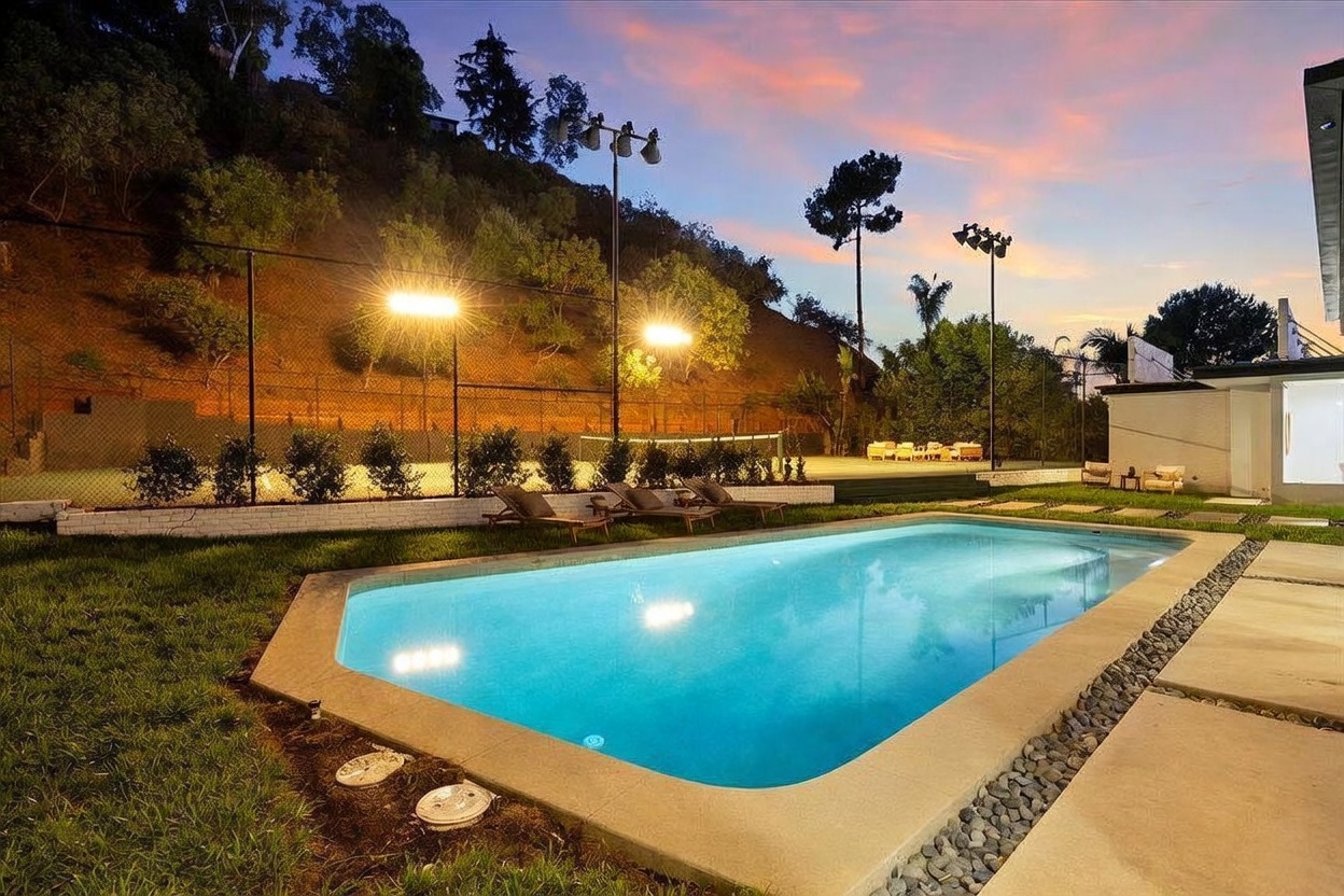 This backyard scene features a rectangular swimming pool set against a backdrop of a lit tennis court and a hillside at dusk. The area includes lounge chairs on the grass, a paved patio space, and tall stadium-style lighting illuminating the recreational facilities. The warm glow of the lights against the twilight sky creates an inviting and luxurious atmosphere for outdoor entertaining.