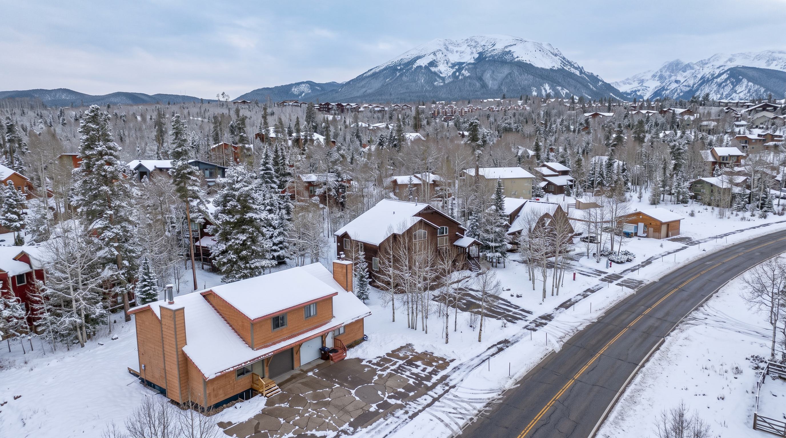 This aerial view captures a rustic, multi-level mountain home surrounded by a snow-covered landscape nestled at the base of a majestic, snow-capped mountain range. The property features wood siding, a large driveway, and convenient side-by-side garages, perfectly situated in a tranquil, pine-filled residential community. The scene exudes a peaceful, alpine aesthetic that highlights both the home's architecture and its scenic natural environment.