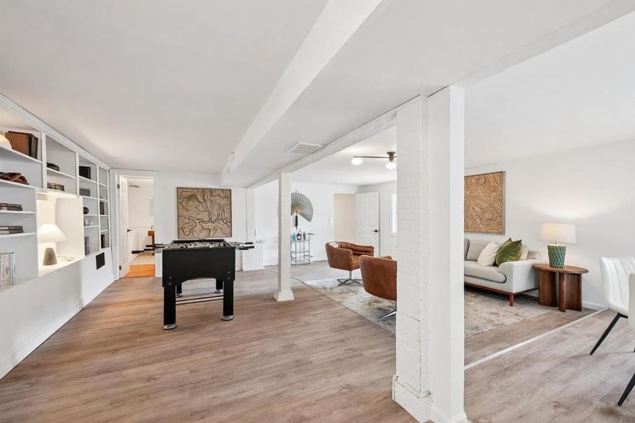 This spacious, open-concept basement features light wood-look flooring and white-painted walls, creating a bright and airy atmosphere. The area is divided into a game zone with a foosball table and a comfortable lounge space furnished with two leather armchairs and a sofa. Structural white-painted brick columns add architectural interest, while built-in shelving provides ample storage and display space.