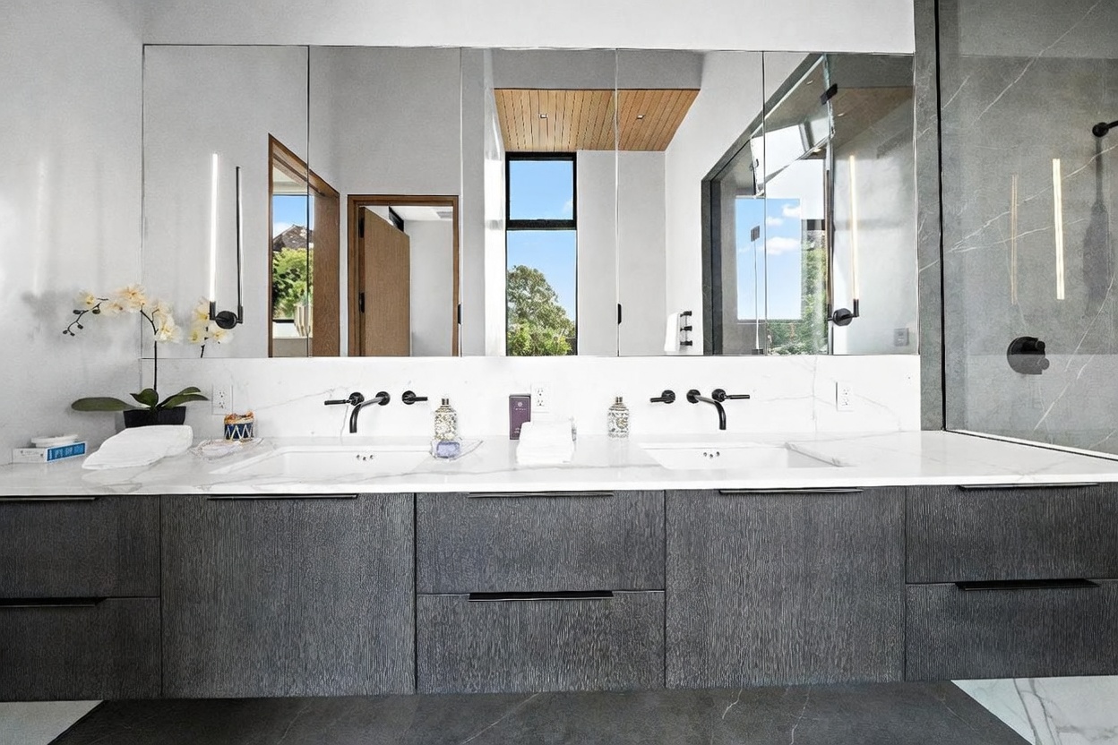 This modern primary bathroom features a dual-sink vanity with dark, textured cabinetry and a white stone countertop. A large, multi-panel mirror spans the wall above the vanity, reflecting a bright, airy space with a tall window and wood-paneled ceiling detail. The clean lines and minimalist fixtures create a sophisticated, contemporary aesthetic.