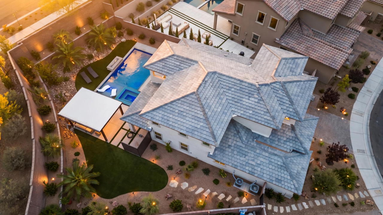 This high-angle aerial perspective captures a modern residential property featuring a sprawling tiled roof and a meticulously landscaped backyard. The outdoor space includes a vibrant blue swimming pool with an integrated spa, a covered patio area, and a mix of artificial turf and desert-inspired rock gardens. The scene is illuminated by warm landscape lighting, creating an inviting and sophisticated atmosphere for outdoor living.