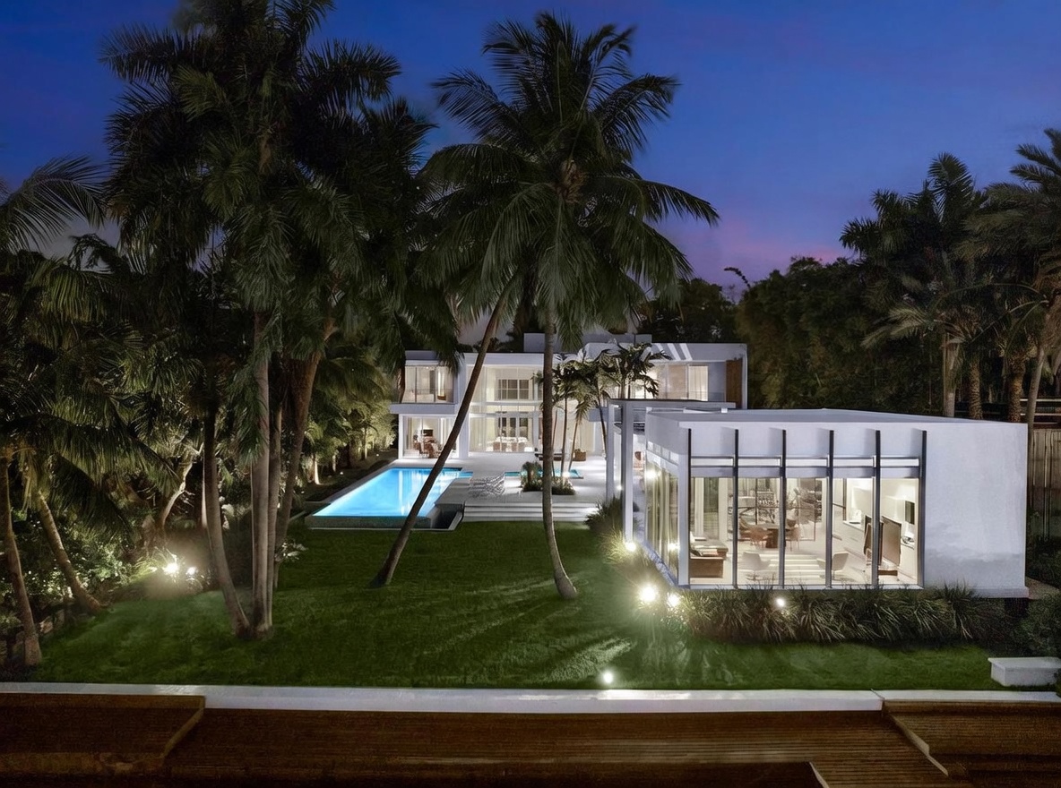 This stunning modern estate is captured at twilight, showcasing a multi-level architectural design with extensive glass walls and clean, geometric lines. The property features a long, rectangular swimming pool set within a lush, manicured lawn framed by tall palm trees and integrated landscape lighting. The perspective is an elevated exterior view, highlighting the seamless transition between the indoor living spaces and the expansive outdoor entertainment area.