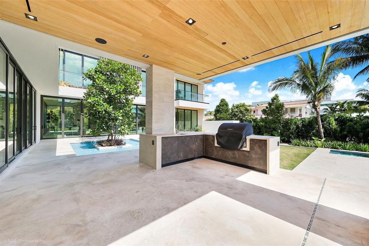 This expansive covered patio features a sleek outdoor kitchen with a built-in grill and stone-patterned cabinetry. The space is defined by a warm wood-paneled ceiling and large glass walls that provide a seamless transition to the interior living areas. A unique water feature with a central tree sits adjacent to the patio, overlooking a lush green lawn and a sparkling pool, creating a sophisticated and tranquil outdoor retreat.