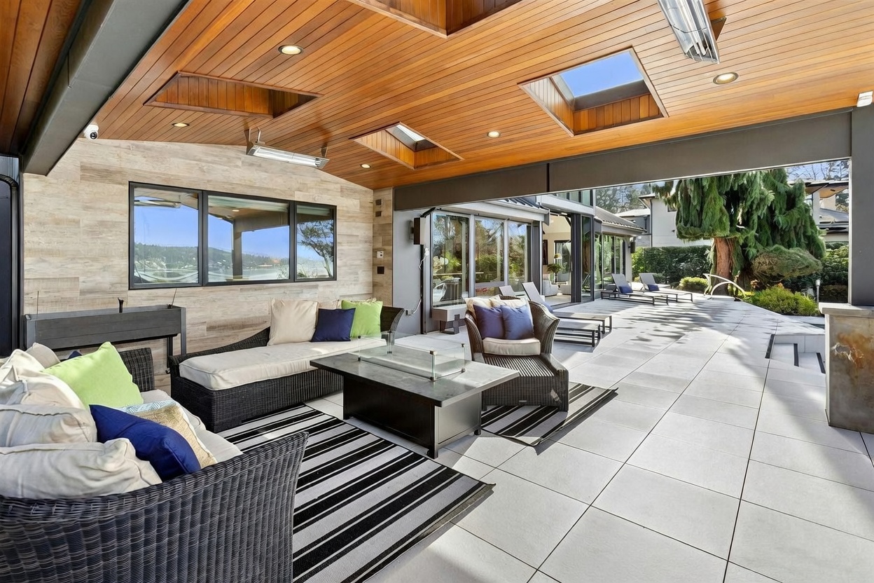This luxurious covered patio features a sophisticated outdoor living space with modern wicker furniture, a central fire pit table, and striped area rugs. The space is defined by a warm wood-paneled ceiling with integrated skylights and recessed lighting, seamlessly transitioning to a spacious tiled terrace overlooking a scenic landscape. The open-air design creates an inviting, high-end atmosphere perfect for entertaining and relaxation.