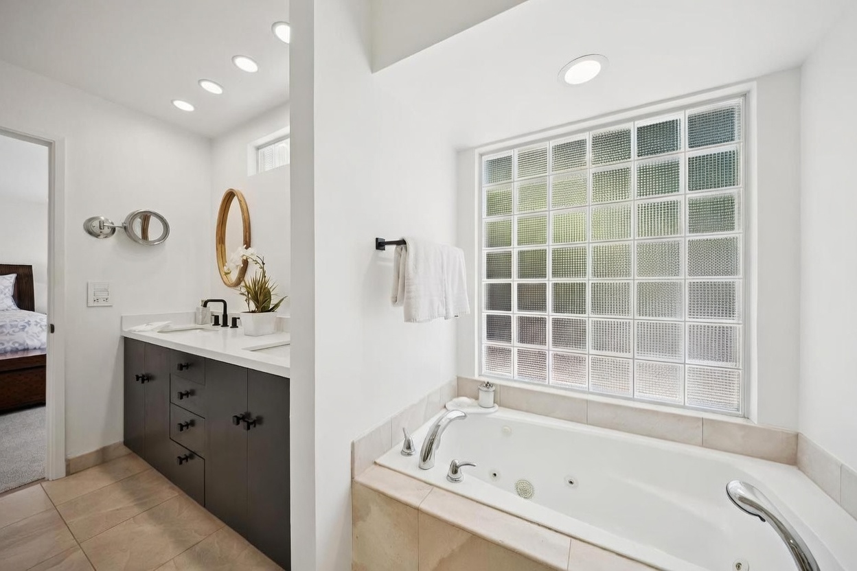 This primary bathroom features a clean, modern aesthetic with a dark-toned vanity topped with a light-colored counter and a circular mirror. To the right, a jetted soaking tub is positioned beneath a large glass block window that provides natural light while maintaining privacy. The space is finished with neutral floor tiles and a wall-mounted towel rack, creating a bright and functional atmosphere.