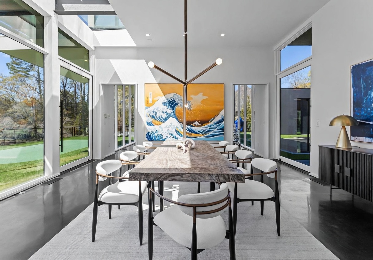 This sophisticated dining room features a live-edge wood table surrounded by modern white chairs, set beneath a striking geometric chandelier. The space is defined by expansive floor-to-ceiling windows that offer abundant natural light and views of the surrounding greenery. A large, vibrant piece of art depicting a stylized wave serves as a bold focal point on the back wall, enhancing the room's contemporary and airy aesthetic.