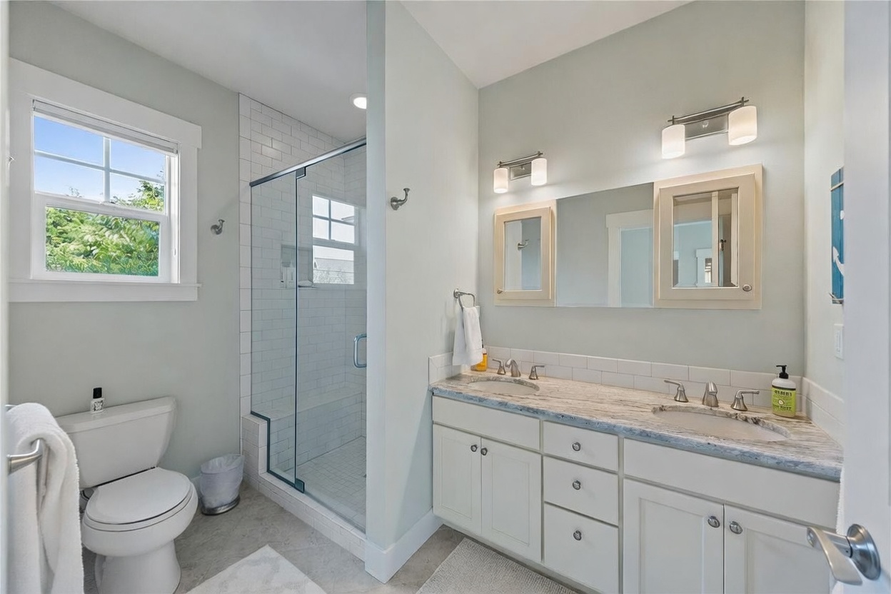 This bright and clean primary bathroom features a dual-sink vanity with a light-toned stone countertop and white cabinetry. A glass-enclosed shower with white subway tile walls and a tiled floor sits adjacent to a standard toilet. The space is illuminated by wall-mounted light fixtures above a large, multi-paneled mirror, creating a functional and airy atmosphere.
