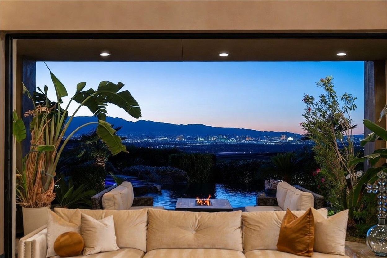 This luxurious indoor-outdoor living space features a plush, neutral-toned sectional sofa facing a stunning, unobstructed panoramic view of a city skyline at dusk. The room seamlessly transitions to a patio area containing a fire feature and a pool, framed by lush tropical greenery and large potted plants. The perspective is from inside the home, looking out through a wide, open aperture that emphasizes the dramatic contrast between the warm, inviting interior and the cool, expansive evening horizon.