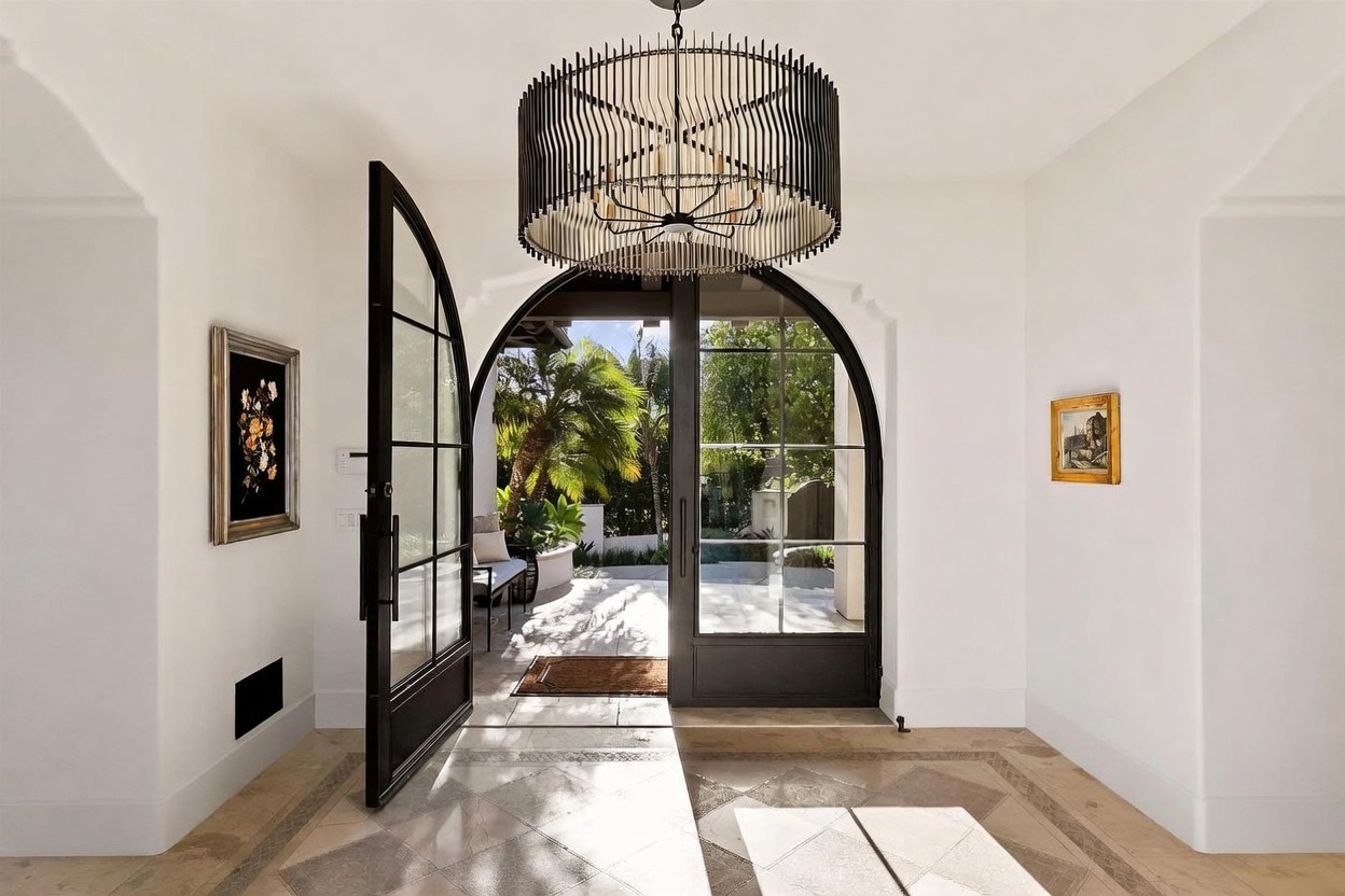 This elegant entryway features a dramatic, large-scale black metal drum chandelier suspended from a high ceiling. The space is anchored by a grand, arched black-framed glass door that opens to a sunlit outdoor patio, creating a seamless transition between indoor and outdoor living. The neutral walls are adorned with framed artwork, and the floor is finished with light-toned stone tiles featuring a decorative inlay border.