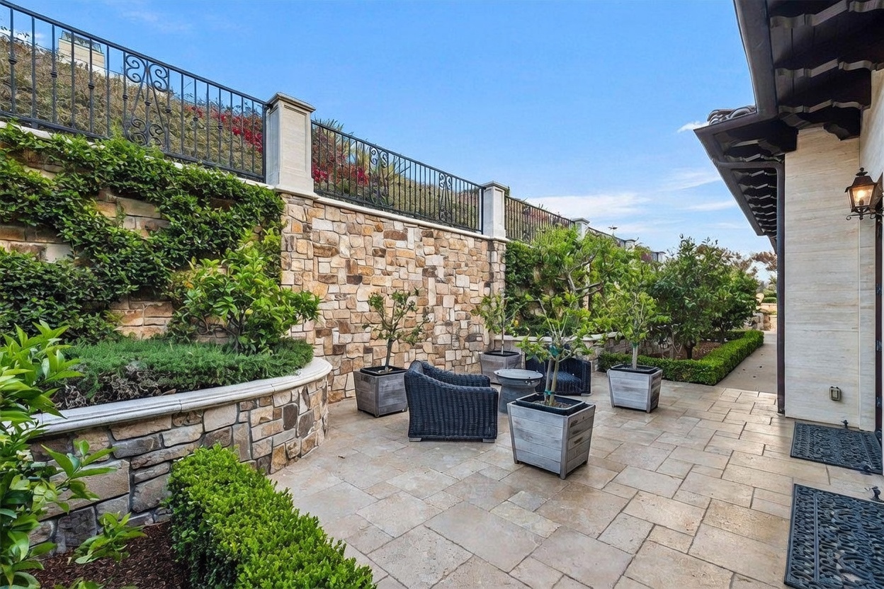 This elegant stone-paved patio offers a serene outdoor living space, featuring a curved retaining wall adorned with lush greenery and climbing vines. The area is furnished with dark wicker seating and potted trees, creating a sophisticated and private retreat. A tall stone wall with decorative iron fencing provides a sense of enclosure, while the adjacent building exterior adds a touch of architectural character.
