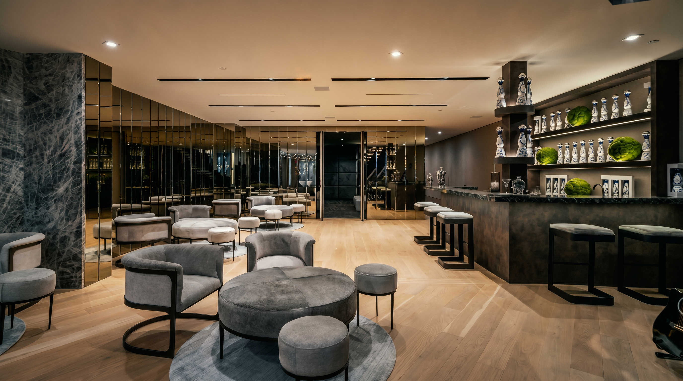 This sophisticated lounge area features a contemporary bar with illuminated shelving and a plush seating arrangement centered around a circular ottoman. The space is defined by its sleek, modern aesthetic, highlighted by mirrored walls, recessed linear lighting, and warm hardwood flooring. The perspective captures the entire room, showcasing a luxurious entertainment space perfect for hosting guests.