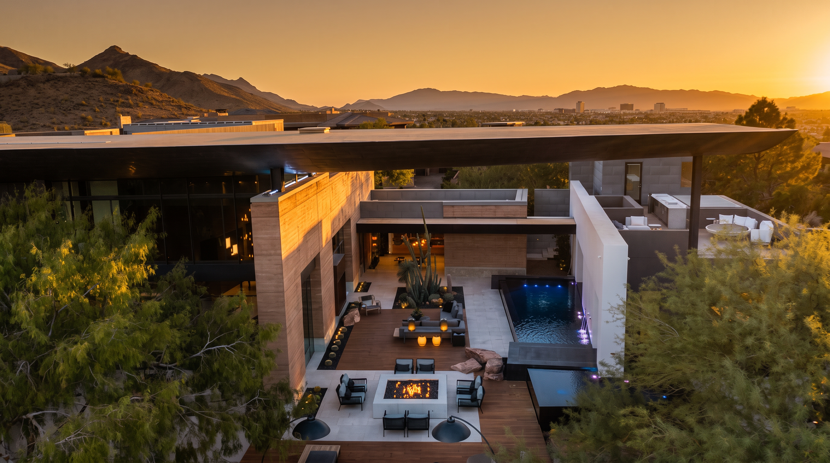 This high-angle aerial view captures a luxurious modern desert estate at dusk, showcasing a seamless integration between indoor and outdoor living spaces. The property features a multi-level deck with a fire pit lounge, a sleek rectangular pool, and a covered patio area, all set against a backdrop of rugged mountains and a distant city skyline. The architecture emphasizes clean lines, expansive glass walls, and warm, earthy tones that complement the surrounding arid landscape.