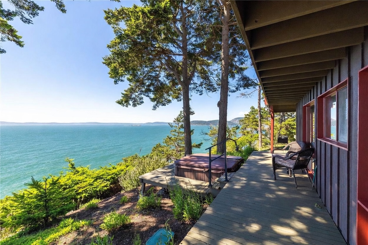 This image captures a spacious wooden deck overlooking a vast, serene body of water with distant hills on the horizon. The deck features a built-in hot tub, comfortable outdoor seating, and a covered section along the side of the house, which is finished in dark-toned siding with red window frames. The perspective is from the deck looking outward, emphasizing the tranquil coastal setting and the seamless integration of the home with its natural surroundings.