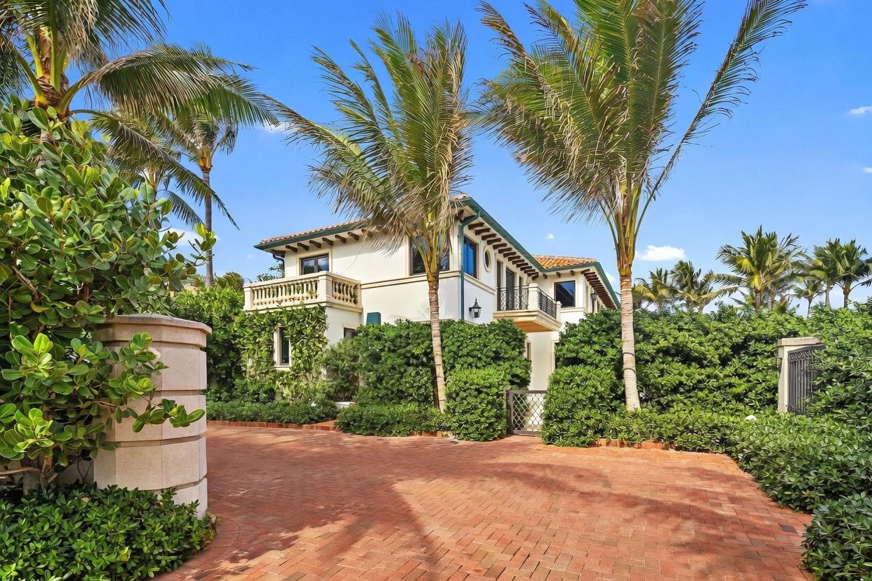 This Mediterranean-style estate features a multi-level facade with a terracotta tile roof and a prominent balcony with decorative railings. The property is accessed via a wide, red brick-paved driveway framed by lush, manicured hedges and tall palm trees. The bright, sunny atmosphere highlights the architectural details and the private, gated entrance to the residence.