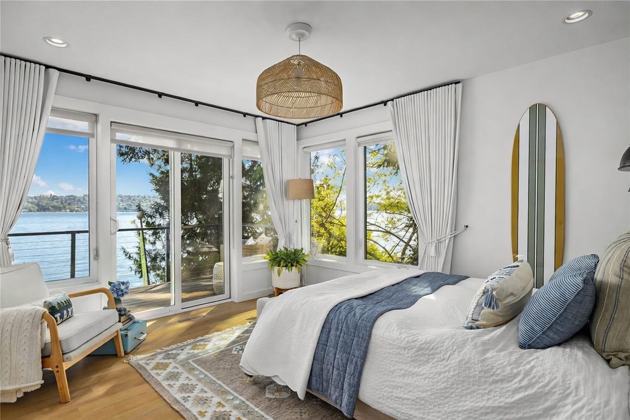 This bright and airy primary bedroom features a large bed with a blue and white color palette, positioned to take advantage of expansive water views through large windows and sliding glass doors. A woven pendant light fixture hangs from the ceiling, adding a natural, coastal texture to the room, while a surfboard mounted on the wall enhances the relaxed, waterfront aesthetic. The space is finished with light wood flooring and a patterned area rug, creating a serene and inviting atmosphere.